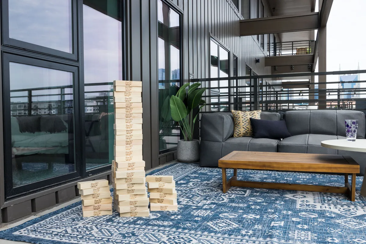 A close-up view of an outdoor sofa and coffee table, with a decorative rug underfoot and a stack of giant Jenga blocks in the center, hints at playful moments. The relaxed, open-air setting suggests a space for games, casual drinks, and laughter, with greenery adding an inviting touch.