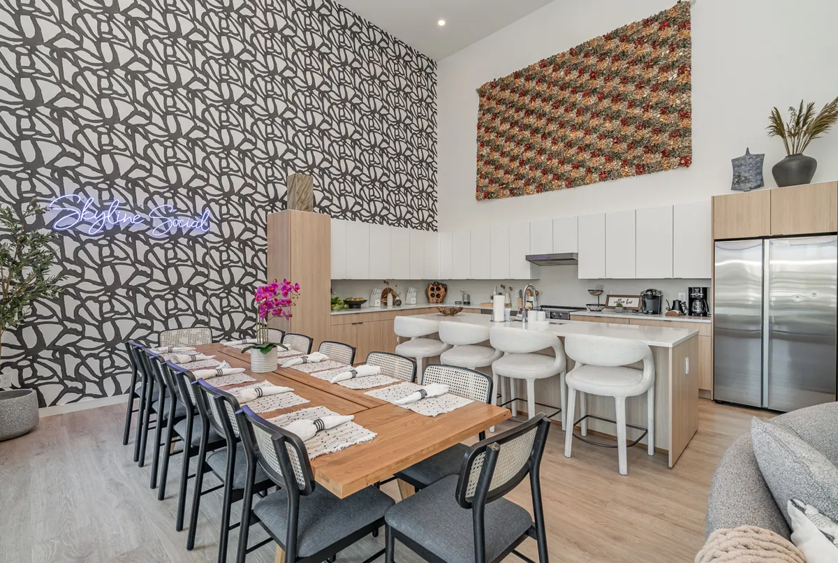 A long wooden dining table set for a meal sits beneath a vibrant patterned wall and a neon 'Skyline Social' sign. Colorful textile art and modern decor create a welcoming, energetic atmosphere, ideal for group dinners and festive gatherings.
