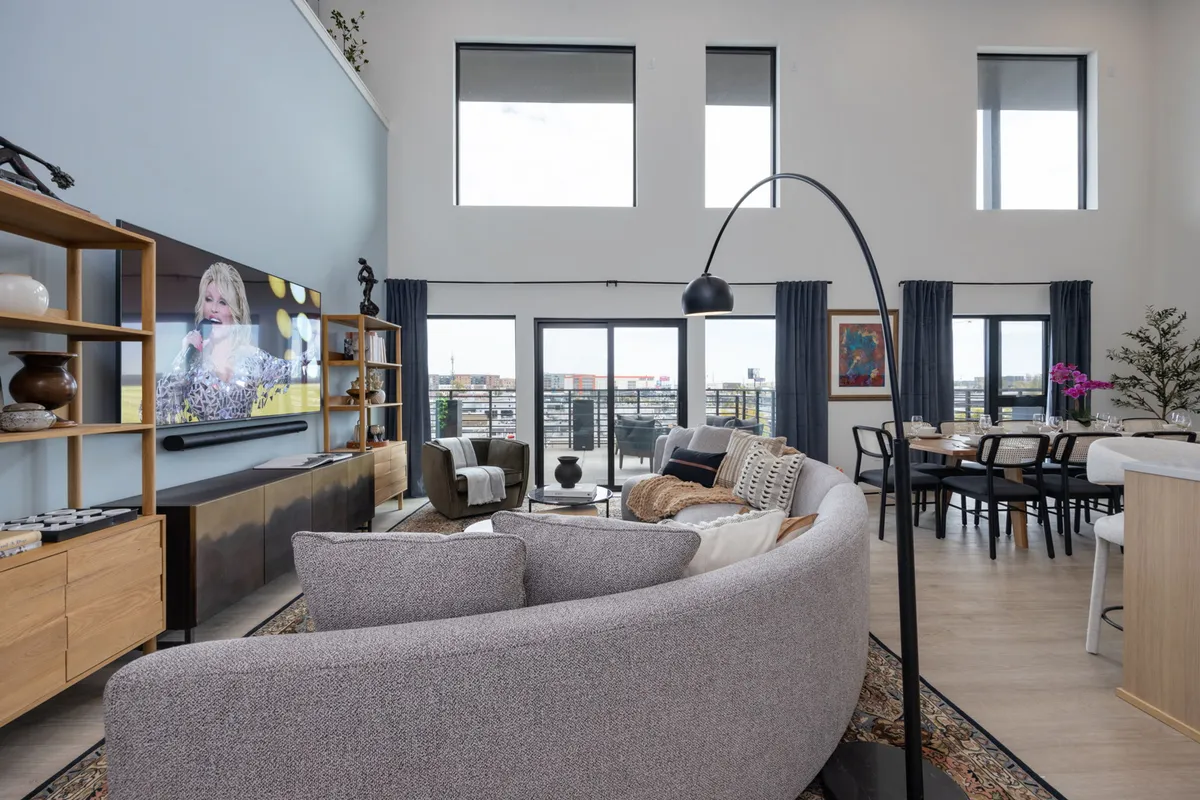 A spacious, open-concept living and dining area is filled with natural light from large windows. A curved sofa faces a large television, while a stylish dining table sits nearby, creating a seamless flow between lounging, dining, and entertainment. The room exudes comfort and sociability, enhanced by modern decor.