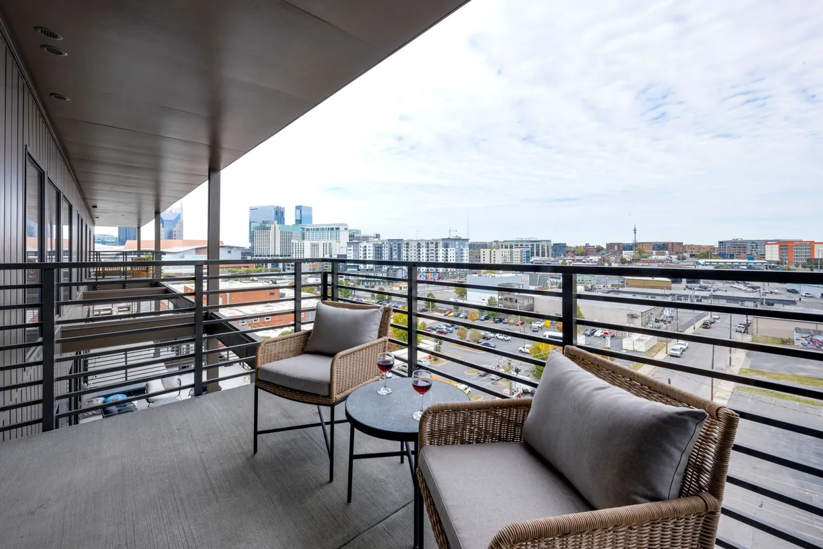 This exterior image features a private balcony seating area overlooking the Nashville cityscape. Two wine glasses rest on the table, hinting at leisurely mornings or sunset drinks, with the skyline providing a dramatic urban backdrop.