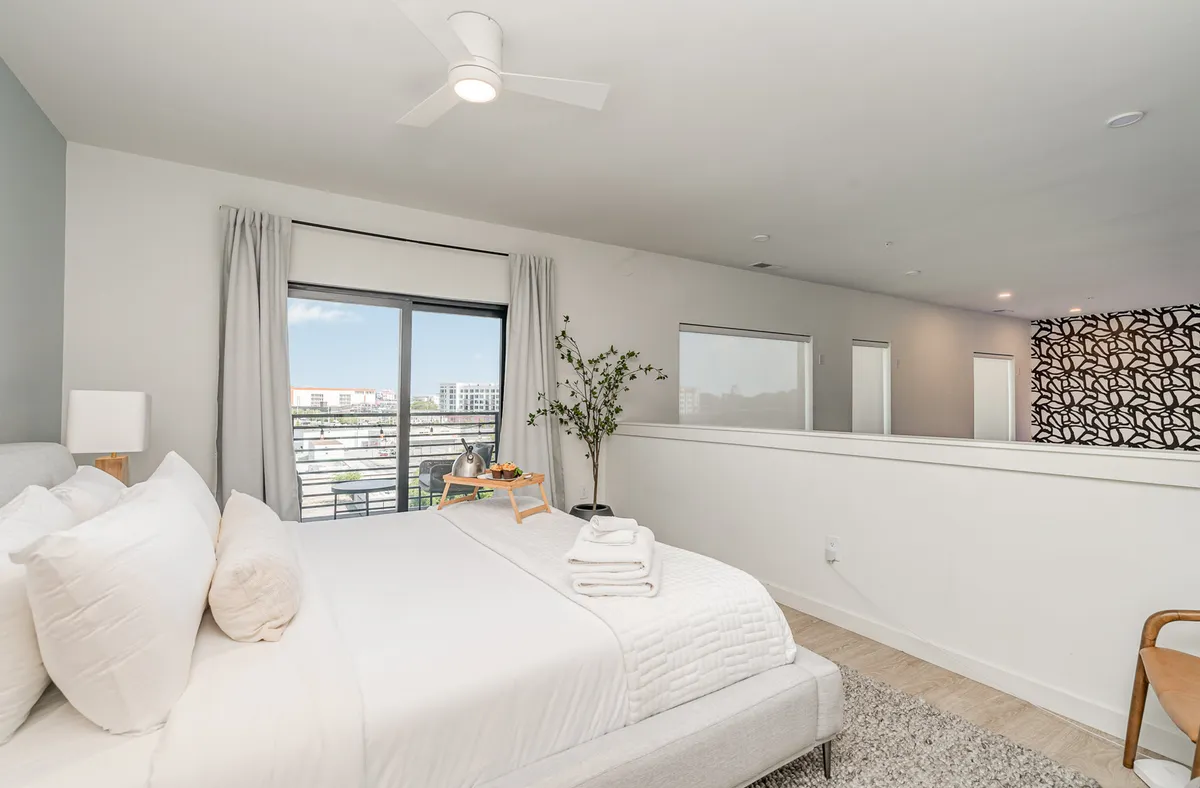 A serene bedroom space with a bed dressed in white linens, large windows letting in abundant natural light, a decorative plant, a modern ceiling fan, and a stylish accent wall. The atmosphere is peaceful and cozy, ideal for restful nights.