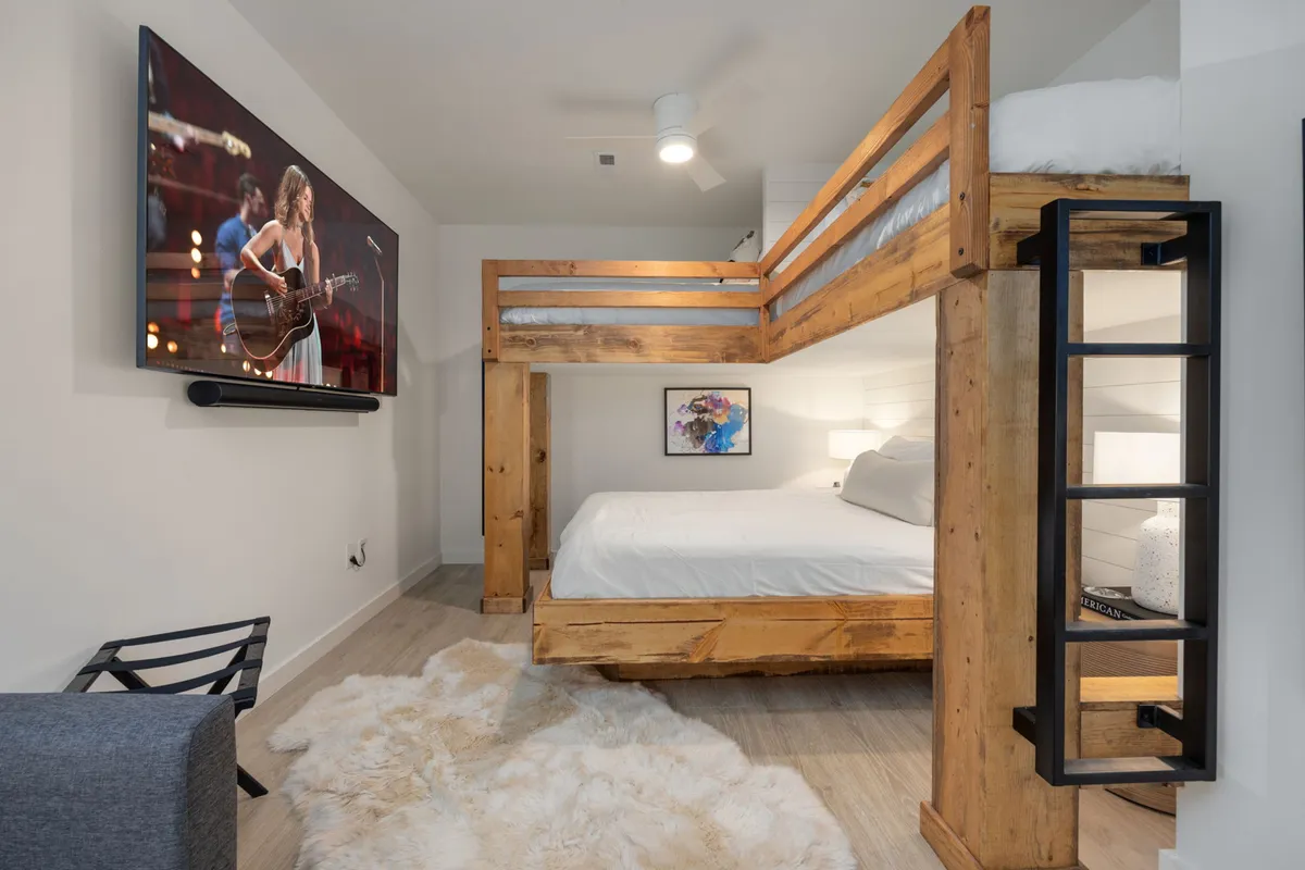 A playful and comfortable bunk room with a twin-over-queen bunk bed, a television mounted on the wall, an area rug for lounging or play, and artwork adding a lively touch to the space.
