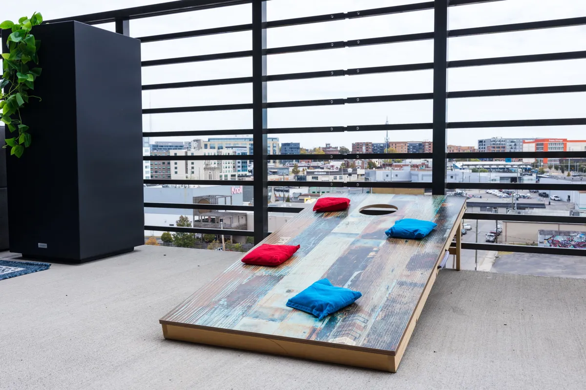 A playful outdoor patio scene with a cornhole game board, colorful bean bags, planters, and an urban background—inviting groups to socialize and enjoy games together.