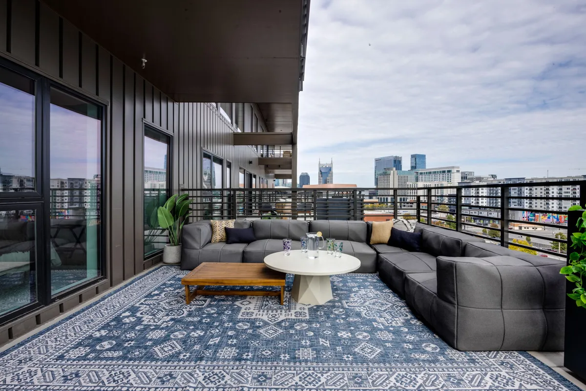 A stylish rooftop lounge furnished with a large sectional sofa, coffee table, and outdoor rug, overlooking the city skyline. The scene is set for relaxing, entertaining guests, or enjoying drinks while watching the sunset over the city.