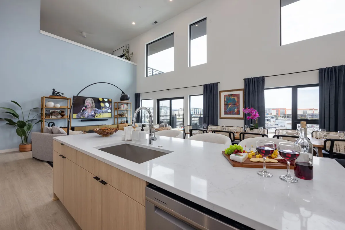 A modern kitchen with a large island opens into a bright living and dining area. A TV is visible, and the space is lively yet relaxed, with barstools at the island and floor-to-ceiling windows letting in abundant light. The setup encourages both cooking and casual socializing.