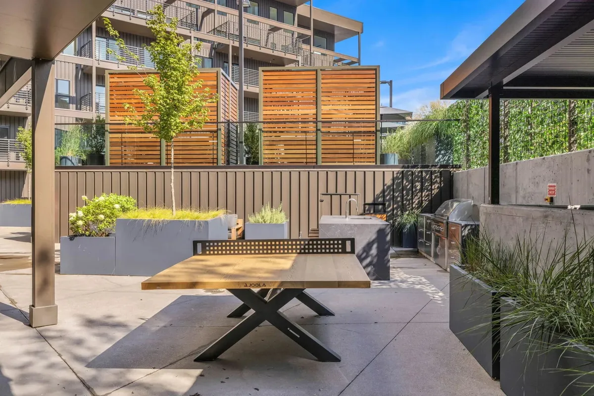 A community BBQ and outdoor dining area with a wooden picnic table, decorative wooden screens, planter boxes with greenery, and a dedicated grill space. The mood is casual and friendly, perfect for group dining and sunset meals.