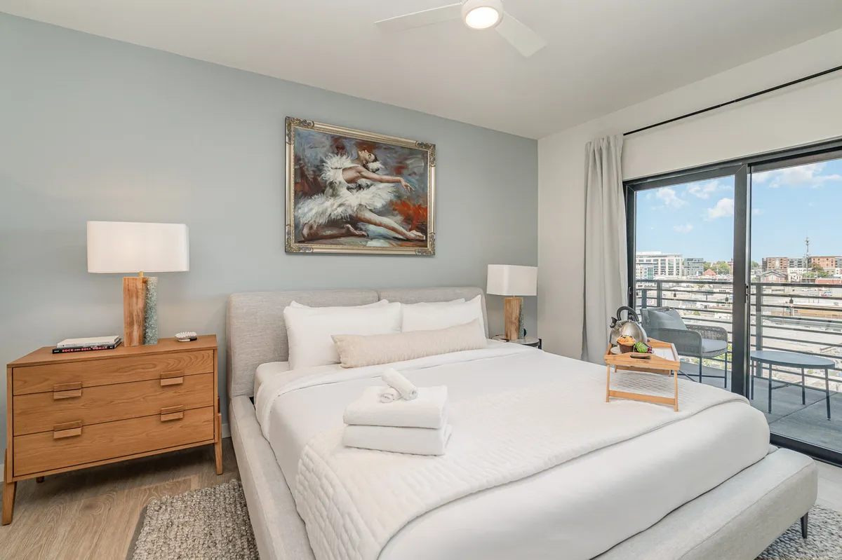 A tranquil bedroom featuring a bed with crisp white linens, a framed painting above the headboard, a nightstand with a lamp, and a private balcony with a view of the city skyline. The room is filled with natural light and exudes a sense of comfort and relaxation.