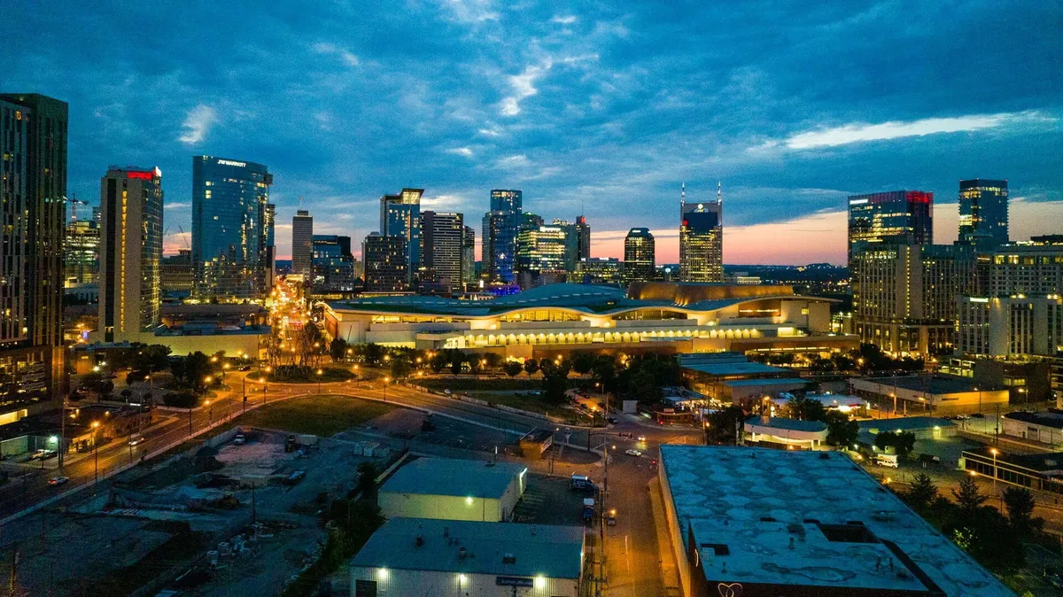 A sweeping view of Nashville's illuminated skyline at dusk, with prominent buildings rising against a dramatic colored sky. Roadways and city lights suggest vibrant nightlife and easy access to cultural hotspots just beyond the residence.