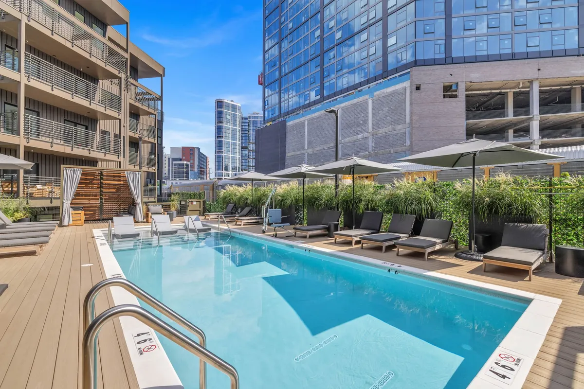 A vibrant exterior shot of a sparkling swimming pool surrounded by lounge chairs and umbrellas, all set within lush, landscaped grounds. The inviting pool area evokes a resort-like experience, perfect for relaxation or social gatherings.